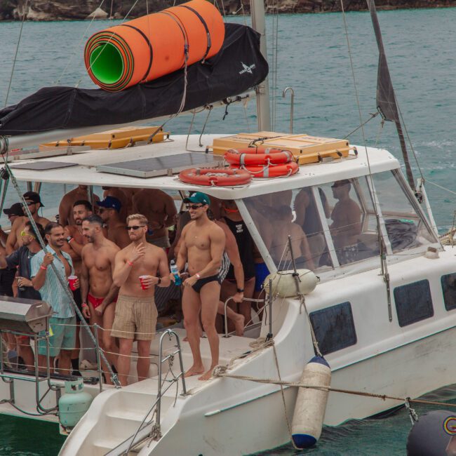 A group of people in swimwear are partying and holding drinks on a white catamaran in the water, with orange floats and black equipment on the boat’s roof. The mood appears lively and festive.