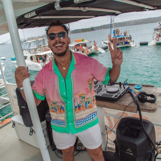 A smiling man in sunglasses and a colorful shirt stands next to DJ equipment on a boat, flashing a peace sign. Other people are partying on nearby boats in the background under cloudy skies.