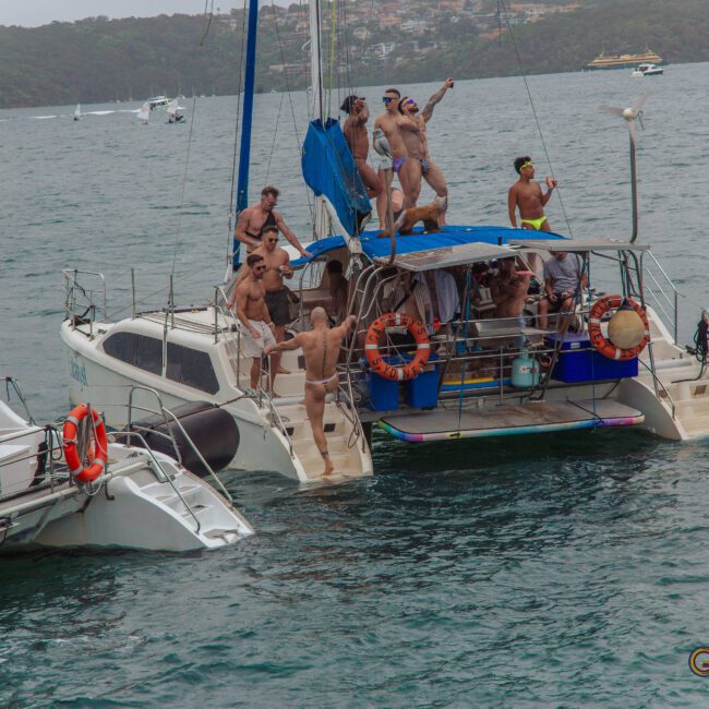 A group of men in swimsuits are partying and dancing on two docked catamarans in open water, with some raising their arms cheerfully and others watching nearby. Trees and buildings are visible on the distant shore.