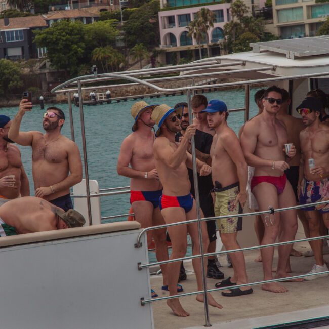 A group of men in swimwear and sunglasses are gathered on a boat, some taking photos and holding drinks, with colorful waterfront houses and trees visible in the background.