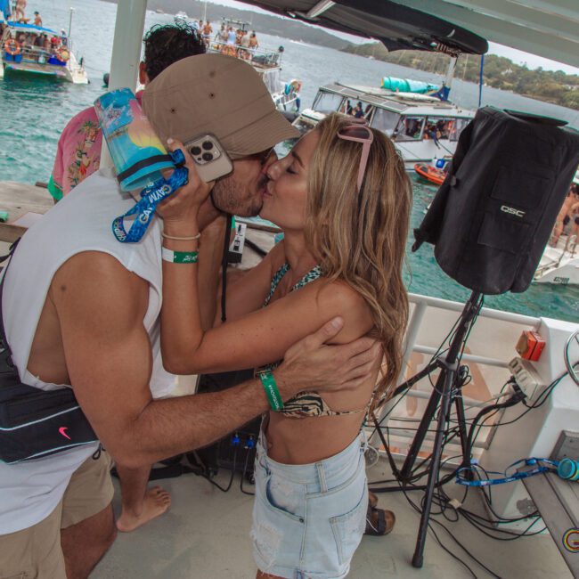 A couple kisses on a boat deck during a lively party, surrounded by music equipment and other partygoers on boats in the water. The atmosphere is festive and summery.
