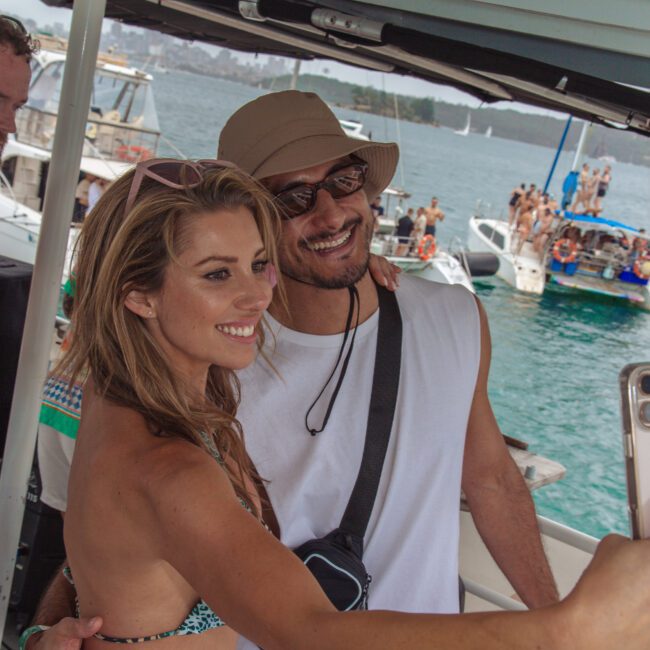 A smiling woman in a bikini and a man in a hat and sleeveless shirt pose for a selfie on a boat, with other boats and people enjoying the water in the background.