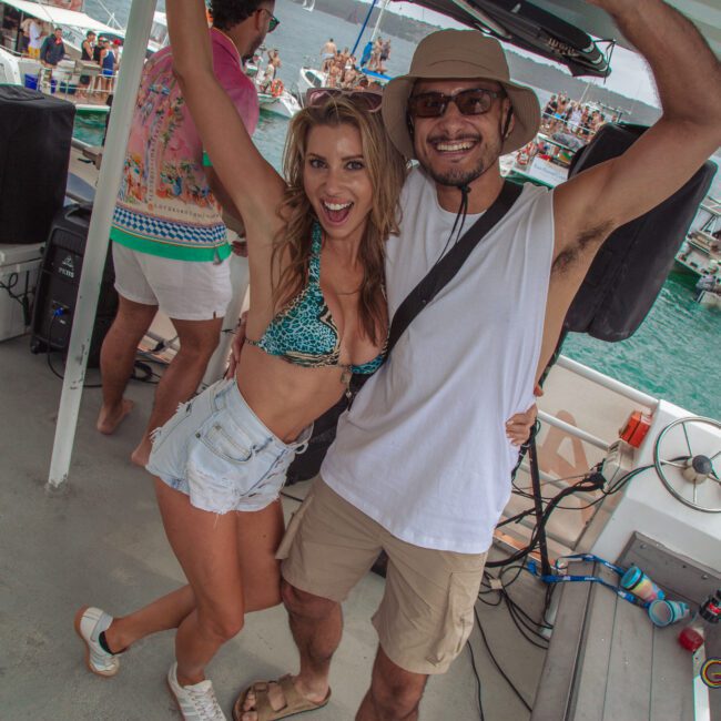 A smiling woman in a bikini top and shorts and a man in a hat and sunglasses pose with arms up on a boat crowded with people, with more boats and a lively party on the water in the background.