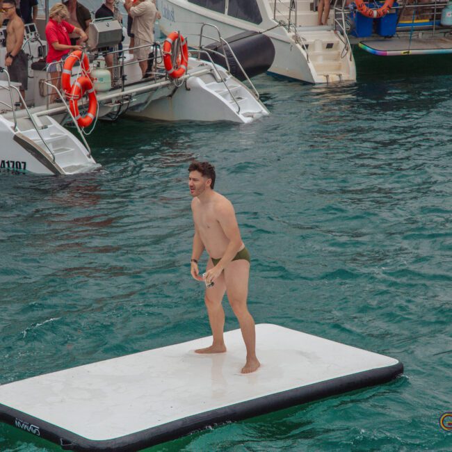 A man in swimwear stands on a floating white mat in the water near boats with people watching and swimming around him. The scene appears lively and fun, suggesting a social or recreational event.