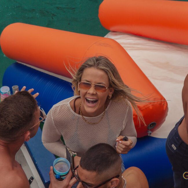 A woman in sunglasses and a mesh top laughs while holding a drink, surrounded by shirtless people on a boat with colorful inflatable floats on the water.