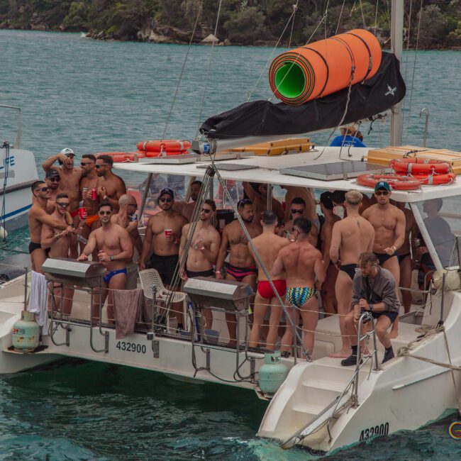 A group of people in swimwear are gathered on the deck of a catamaran, partying and smiling. The boat is on blue water, with another boat nearby and trees along the shore in the background.