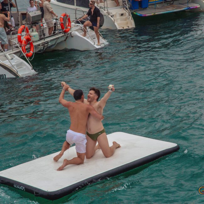 Two men in swimwear kneel, laugh, and hold hands on a floating mat in the ocean, surrounded by people on nearby boats at a lively party. The mood is festive and celebratory.