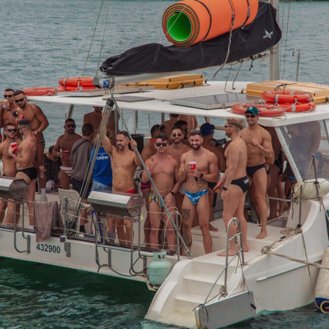 A group of men, mostly shirtless and wearing swimwear, stand and socialize on the deck of a catamaran boat in the water. Some hold drinks and smile, while others talk. The atmosphere appears festive and relaxed.