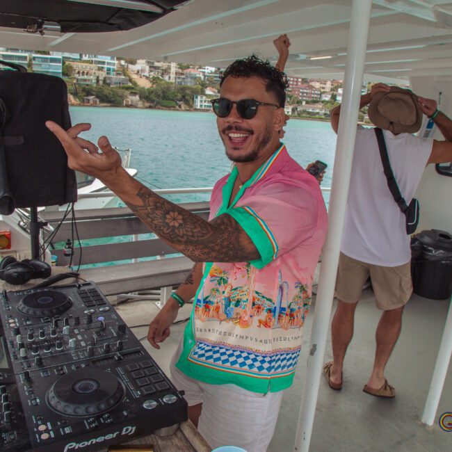 A smiling DJ in a colorful shirt stands at a mixing console on a boat. Another person dances in the background. Water, buildings, and another boat are visible outside. The scene appears lively and festive.