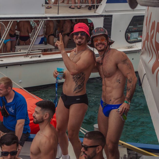 Two men in swimwear pose and smile on a crowded boat party, surrounded by other people in swimsuits. Boats and water are visible in the background, suggesting a festive atmosphere.