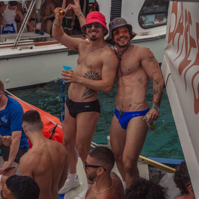 Two men in swimwear and hats pose and smile on a crowded boat during a lively daytime party, surrounded by people in swimsuits holding drinks, with another boat and clear water in the background.