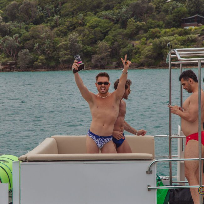 A group of men in swimwear stand on a boat, smiling and having fun. One man in the foreground raises his drink and flashes a peace sign. The boat is on water, with greenery and trees visible in the background.