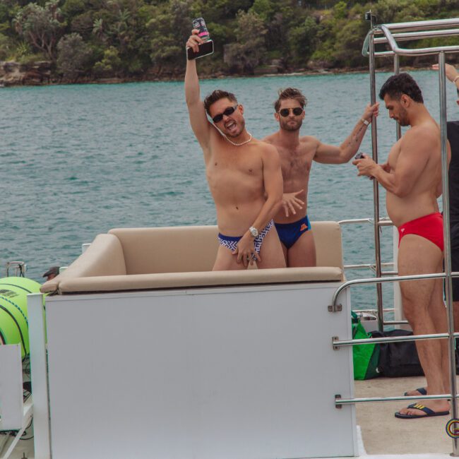 A group of men in swimsuits enjoy a party on a boat by the water; one man in the foreground raises a drink and poses energetically while others stand nearby chatting and holding drinks.
