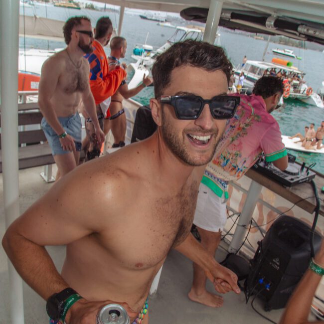 A man wearing sunglasses, a colorful beaded swimsuit, and holding a drink smiles at the camera on a lively boat party, with other people dancing and enjoying themselves in the background near the water.