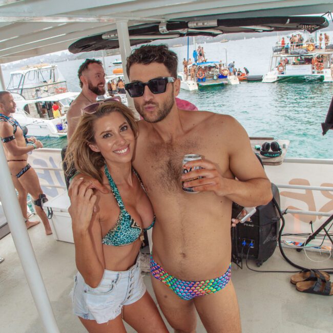 Two people in swimsuits pose and smile for the camera on a boat, with other boats and people in the water behind them. The man is holding a drink, and both look relaxed and festive at a lakeside party.