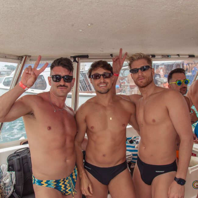 Three men in swimsuits and sunglasses pose on a boat, making peace signs and smiling. Other people in swimwear are in the background, with water and boats visible outside the boat's window.