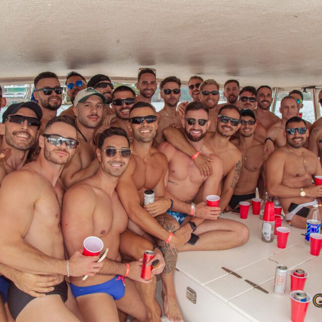 A large group of men in swimwear smile and pose closely together on a boat, holding red cups. Several empty cans and cups are scattered around. The atmosphere appears festive and relaxed.