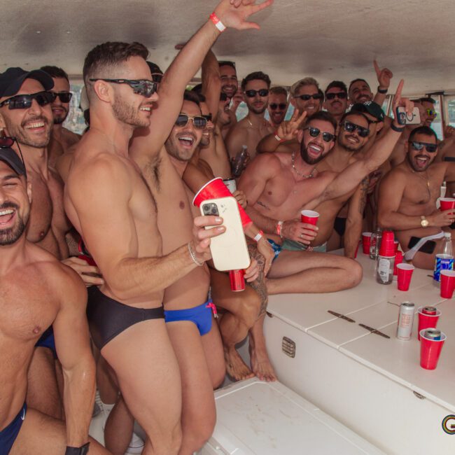 A group of men wearing swimwear smile and pose together on a boat, holding drinks and red cups, appearing to enjoy a lively party atmosphere.