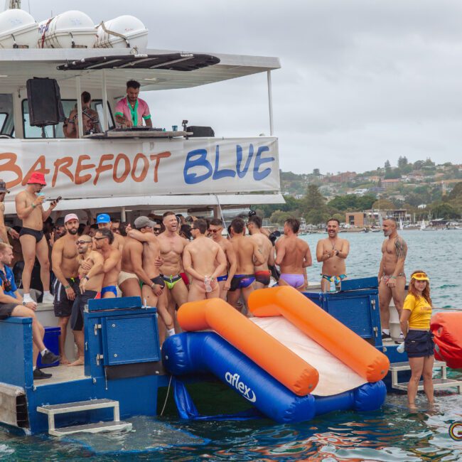 A group of men in swimwear gather and socialize on a boat named "Barefoot Blue," which is anchored on the water. There’s an inflatable slide, and one person in yellow stands near the edge. The shoreline is visible in the background.