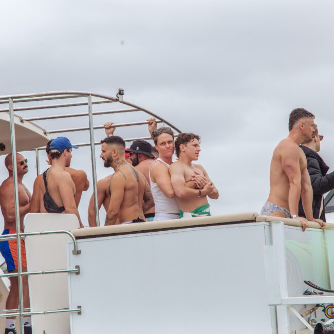 A group of men in swimwear are standing on the deck of a boat under a cloudy sky, some looking out at the water while others chat or pose near the railing.