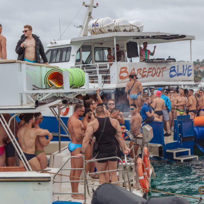 A lively group of people in swimwear gathers on two docked boats, socializing and enjoying the water. The boat named "Barefoot Blue" is visible, with inflatable water activities nearby. The atmosphere is festive and relaxed.