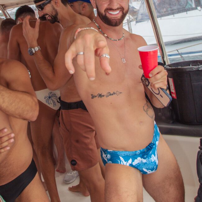 A smiling man in blue swim briefs and a red cup points at the camera on a boat with other shirtless people, enjoying a lively party. A large tiger tattoo is visible on his thigh.