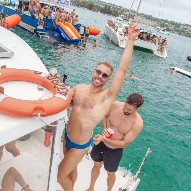 Two men in swimwear stand on the deck of a boat at a lively party on the water. One man poses with an arm raised, smiling at the camera. Other people are seen swimming and socializing on nearby boats.
