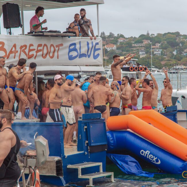 A large group of people in swimsuits enjoy a lively party on a boat called "Barefoot Blue," with music, drinks, and an inflatable slide leading into the water; the background shows houses on a hillside.
