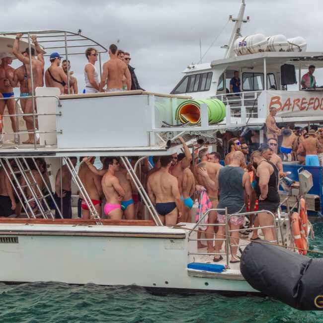 Two crowded party boats are anchored side by side on the water, with many people in swimsuits socializing, standing, and sitting on deck. The atmosphere appears lively and festive under cloudy skies.