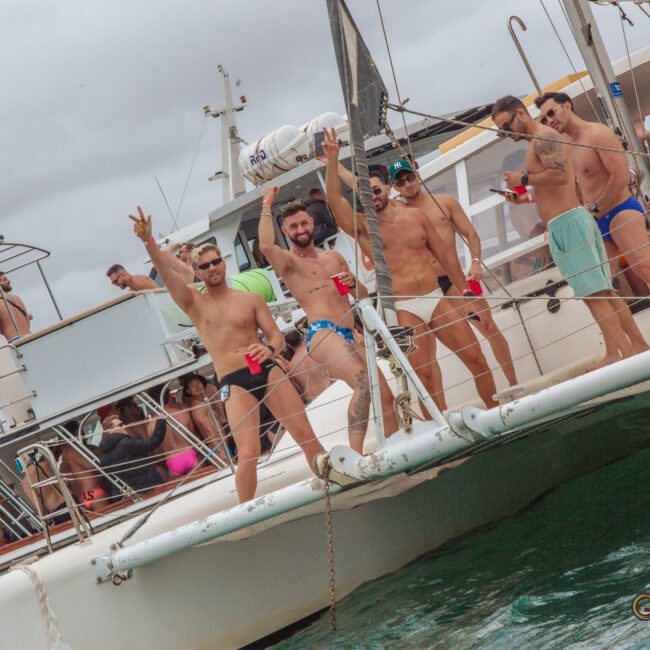 A group of men in swimwear stand on the edge of a boat, smiling and raising drinks, while other people relax on boats nearby. The scene is festive and takes place on the water under a cloudy sky.