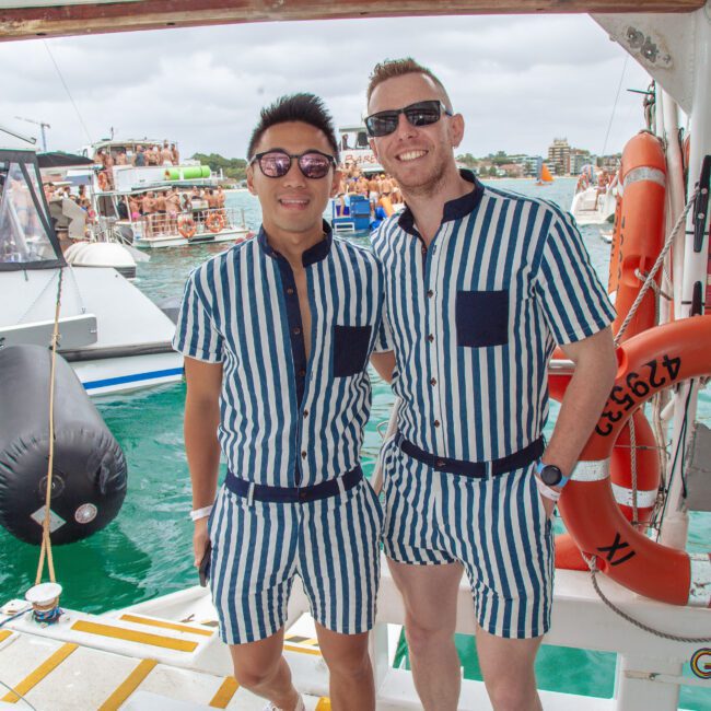 Two men smile on a boat, wearing matching blue and white striped short-sleeve shirts and shorts. Behind them, other boats and people are visible on the water during a lively gathering.
