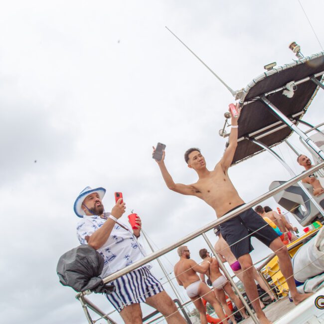 A group of people in swimwear enjoy a party on a boat under cloudy skies. Some are dancing and holding drinks, while others are talking. Towels, bags, and other items are scattered on the deck.