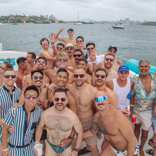 A large group of smiling men in swimwear and summer outfits pose together on a boat, with water and the Sydney Harbour Bridge visible in the background on a cloudy day.