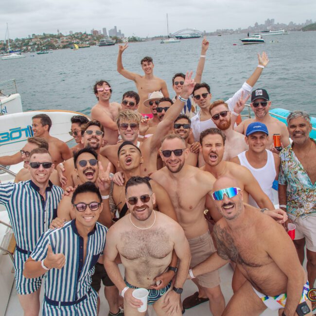 A large group of men in swimwear smiling and posing together on a boat, with water, sailboats, and a city skyline visible in the background under a cloudy sky.