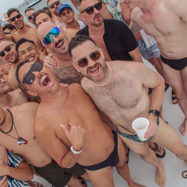 A large group of men in swimwear laugh and pose together on a boat under cloudy skies, enjoying a lively outdoor party. Some wear sunglasses, necklaces, or hats, and most appear to be smiling or cheering.
