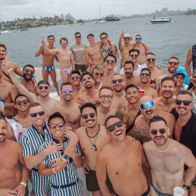 A large group of men wearing swimwear and sunglasses smile and pose together on a boat. The background shows a city skyline, water, and other boats. The mood is festive and joyful, suggesting a fun gathering or party.