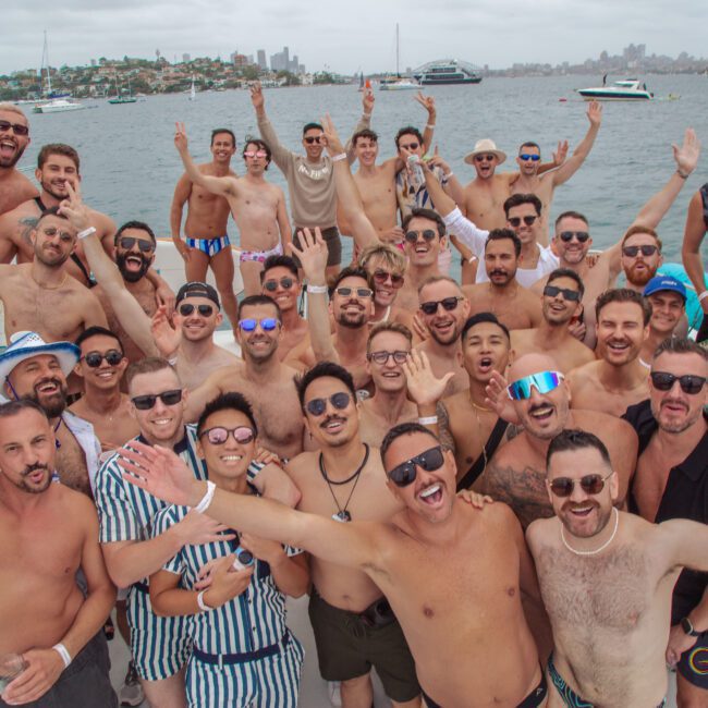 A large group of smiling men pose together on a boat, many shirtless and wearing sunglasses, with arms raised in celebration. The boat is on the water with city buildings and other boats in the background.