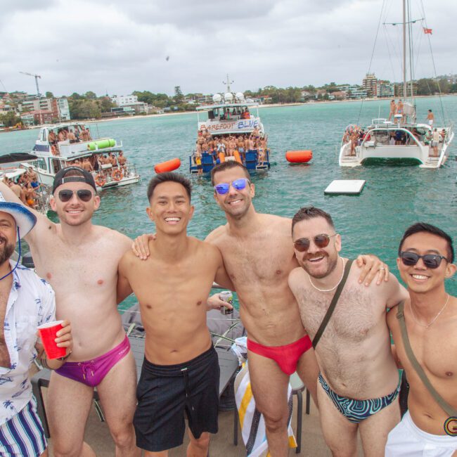 Six smiling men in swimwear pose on a boat with a city shoreline and several crowded party boats in the background, under a cloudy sky. The atmosphere is lively and festive.