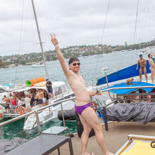 A man in purple swim briefs stands on a boat deck, smiling and making a peace sign. Other people in swimwear are on nearby boats and in the background, with water and houses visible under a cloudy sky.