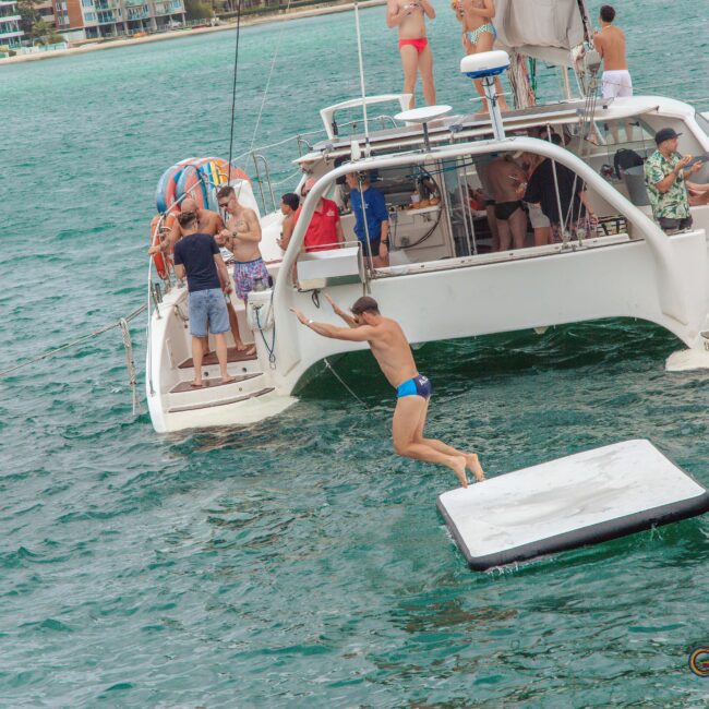 A man in swim trunks jumps from the back of a catamaran onto a floating mat in the ocean, while people on the boat watch. The water is turquoise and there are more boats and buildings in the background.