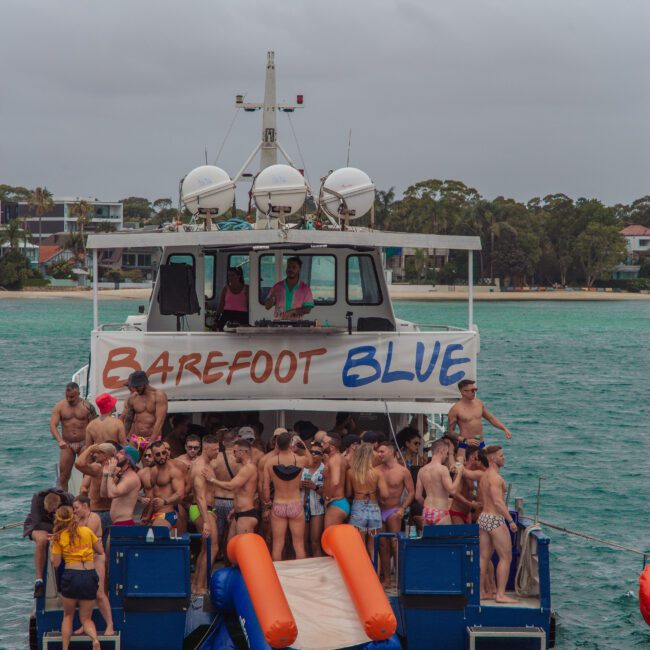 A large group of people in swimsuits gather and socialize on the back deck of a boat named "Barefoot Blue," anchored in turquoise water near a shore with trees and houses under a cloudy sky.