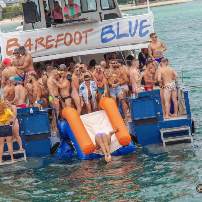 A group of people in swimsuits gather on the back of a boat named "Barefoot Blue," with one person going down an inflatable slide into the water. The scene is festive and energetic, with a tropical shore in the background.