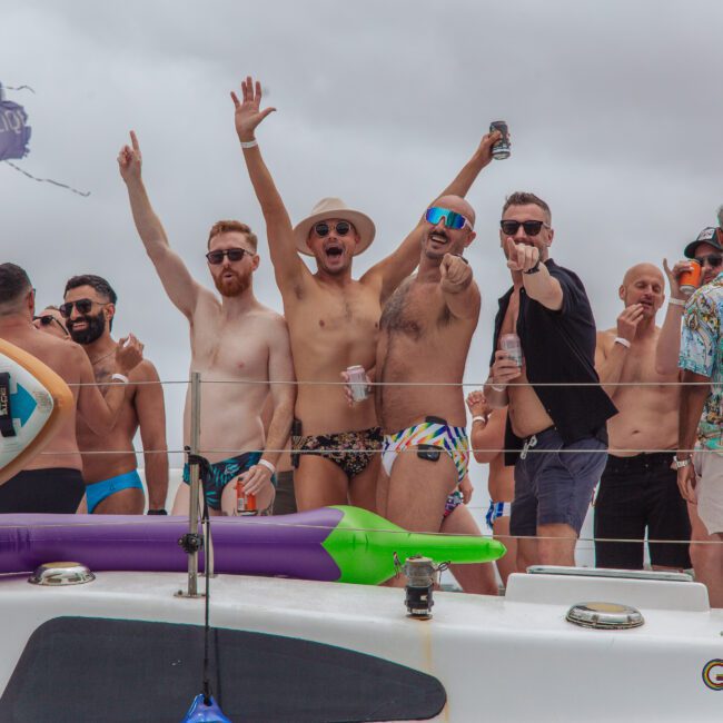A group of men in swimwear stand on a boat, smiling and cheering at the camera. Some are holding drinks and wearing sunglasses. Surfboards and pool floats are visible, with a cloudy sky in the background.