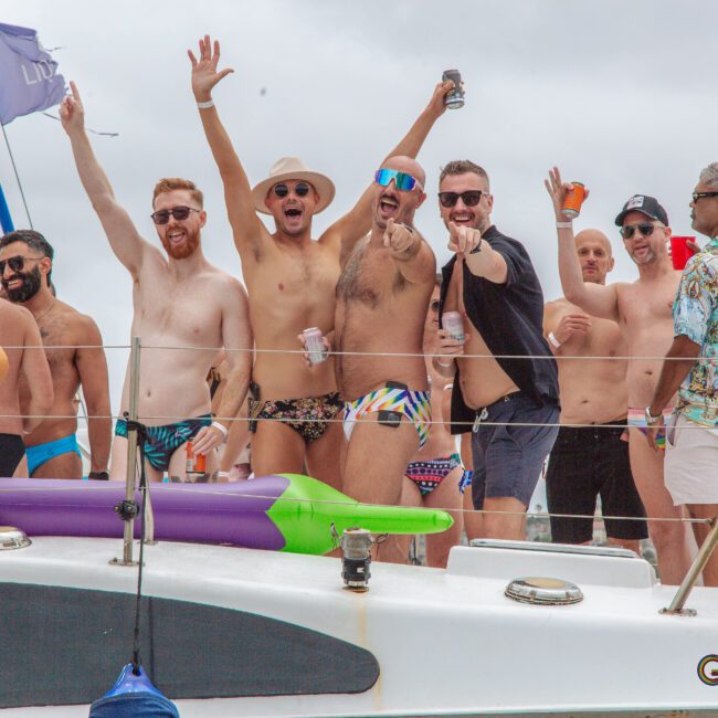 A group of men in swimwear stand and pose cheerfully on a boat, some raising their arms or holding drinks, with pool floats and life rings visible on the deck under a cloudy sky.