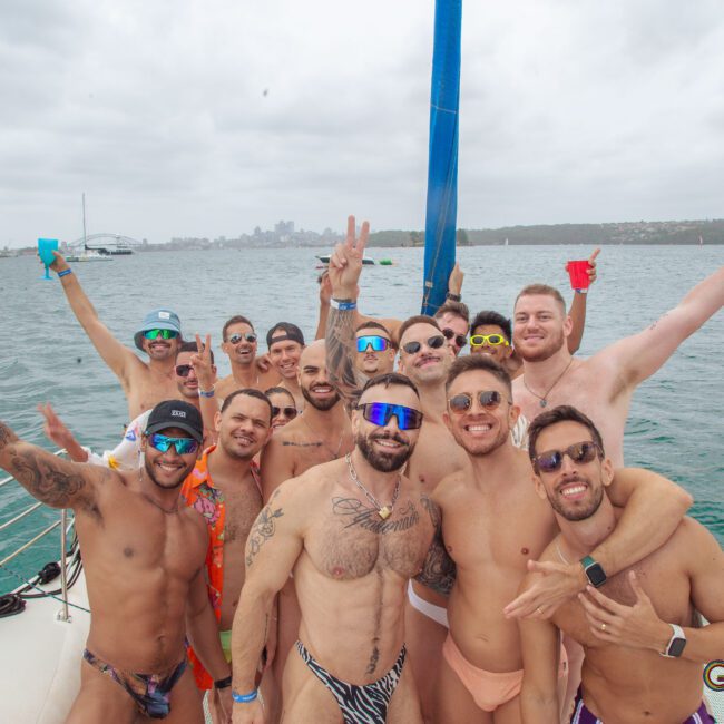 A group of men in swimwear pose and smile on a boat with the city skyline and water in the background under a cloudy sky. Some wear sunglasses and hold drinks, enjoying a festive atmosphere.