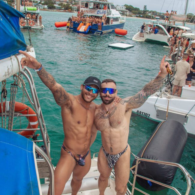 Two men in swimwear smile and pose arm-in-arm on a boat's steps, with a festive crowd on boats in the background on a cloudy day. The scene is lively, with people swimming and socializing in the water.