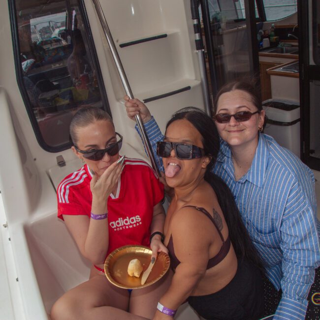 Three women sitting together on a boat, smiling and posing playfully. One woman sticks out her tongue, another covers her mouth while holding a gold plate with food, and the third wears sunglasses and smiles at the camera.