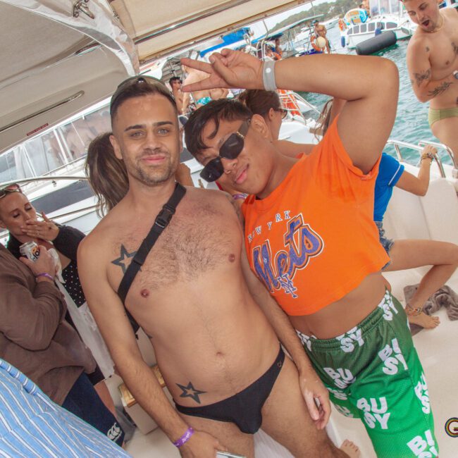 A group of people on a boat party; one person poses in black swimwear with star tattoos, another in green shorts and orange Mets shirt makes a peace sign. Others in swimwear chat and relax with boats and water in the background.