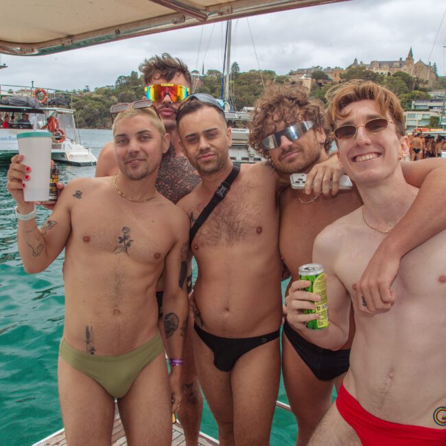 Five men wearing swimwear pose together and smile on a boat with drinks in hand. Other boats and people are visible in the water and onshore in the background, creating a festive summer atmosphere.