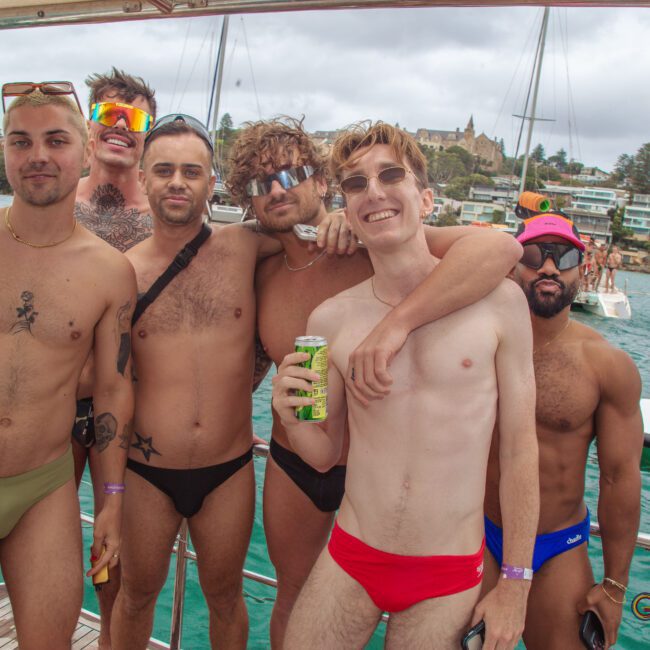 Six men in swimwear stand and pose together on a boat, smiling and holding drinks, with a marina, water, and buildings visible in the background.